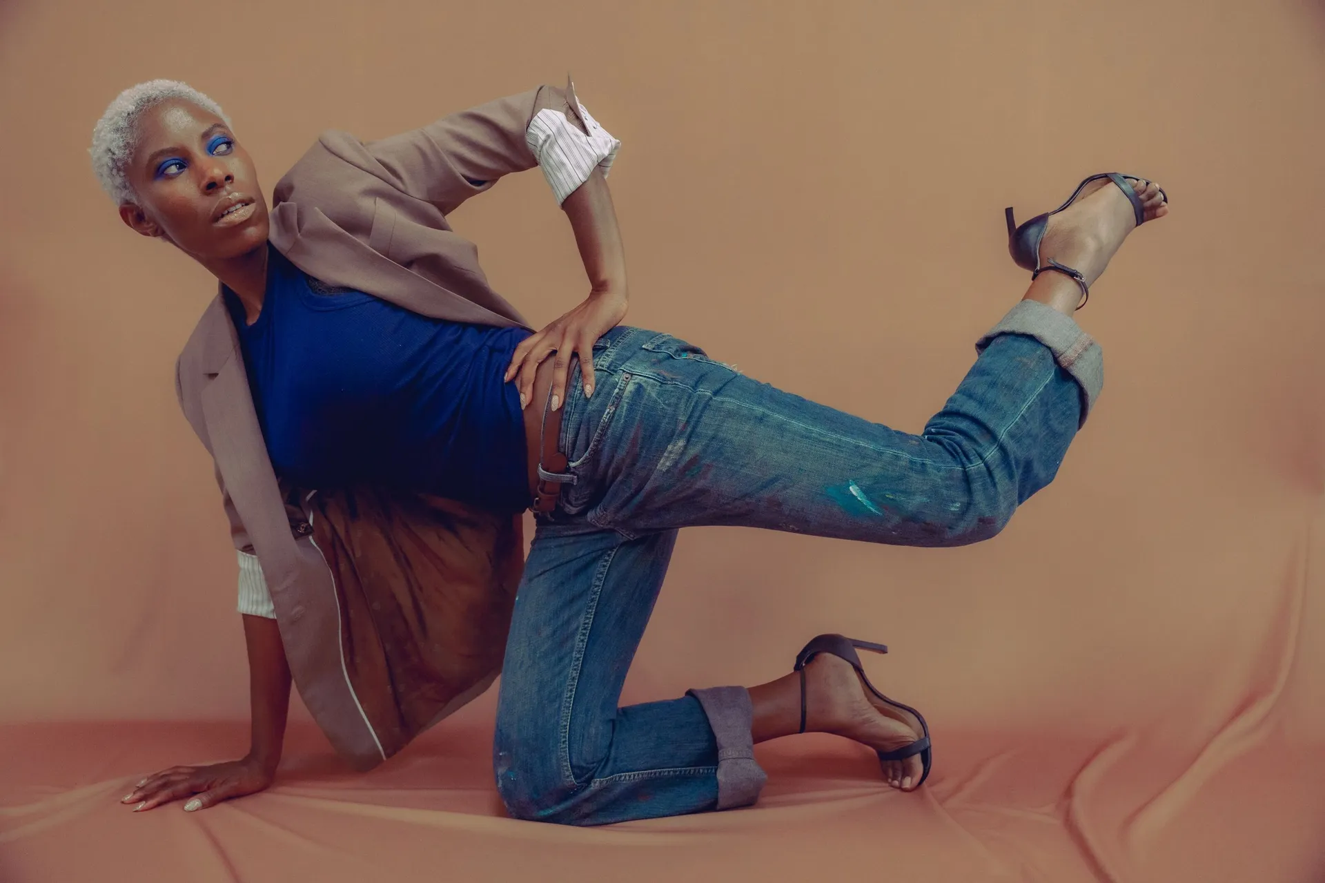 A model in rolled mid-wash jeans and heels strikes a kneeling pose against a tan backdrop